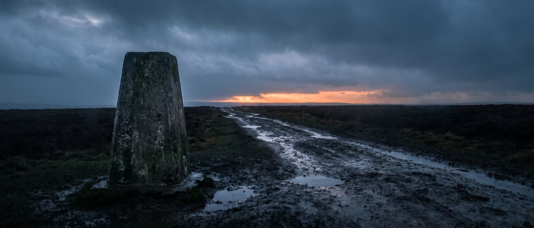 An uncarved stone road marker standing beside an empty track at storm dusk